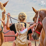 Little girl on her mom's back petting a horse